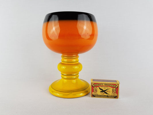 a large orange and black glass goblet with a yellow base, placed next to a matchbox.