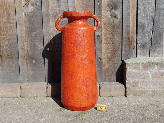 A large, red, ceramic vase with two handles stands against a wooden fence.