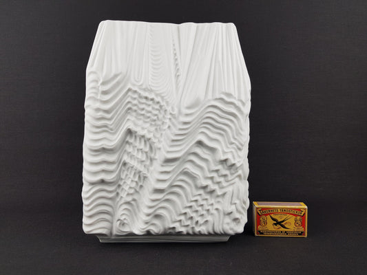 a white, abstract sculpture with a textured, wave-like design, placed next to a matchbox.