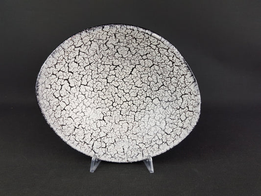 A black and white ceramic plate with a cracked, speckled texture is displayed on a clear stand against a dark background.