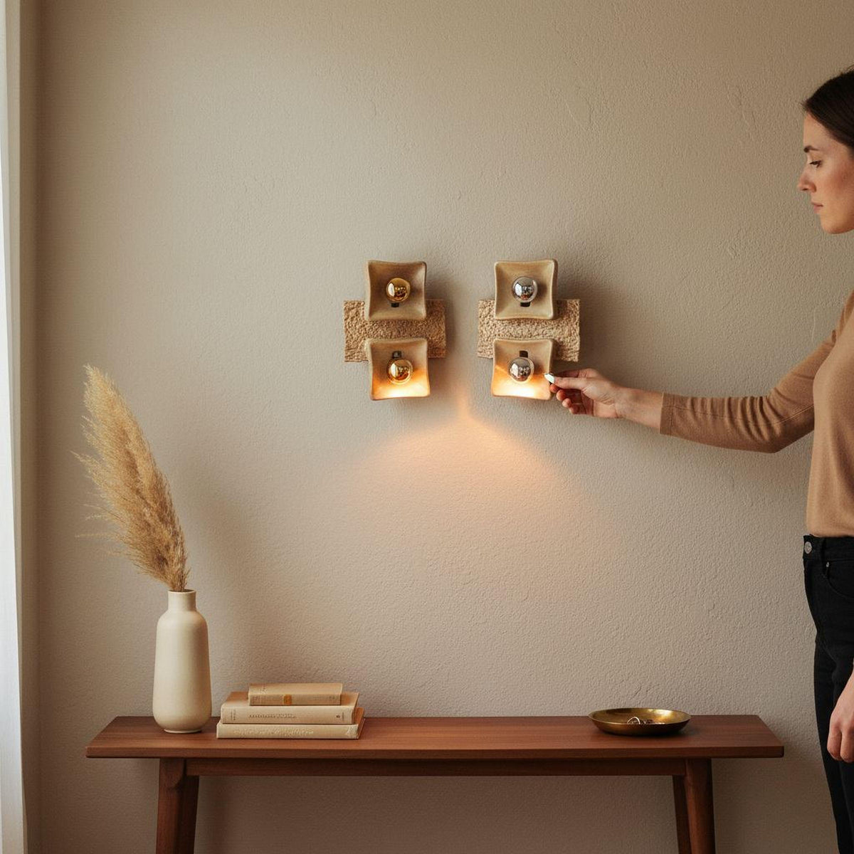 A person is standing next to a wooden console table, which has a vase, books, and a bowl on it. The person is holding a light switch, possibly to turn on or off the lights above the console table.