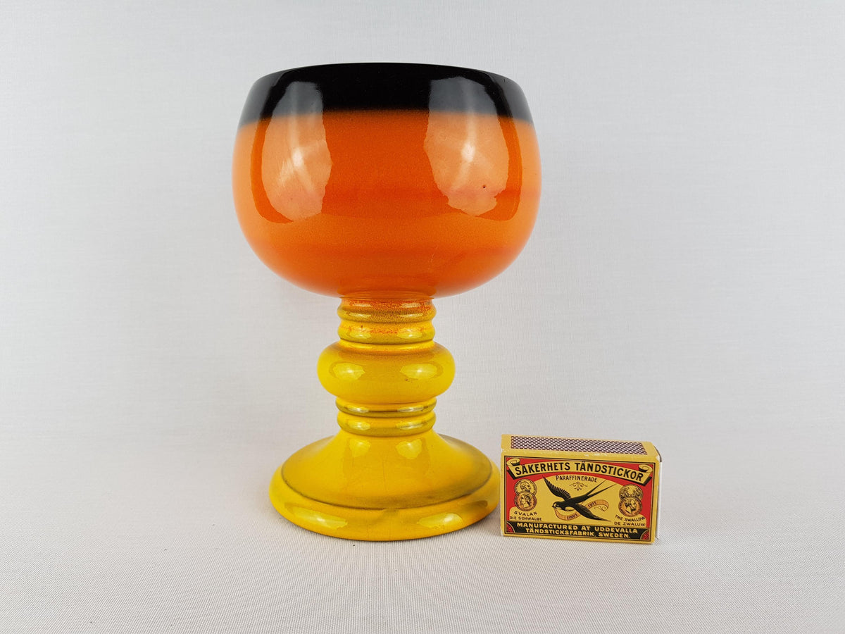 a large orange and black glass goblet with a yellow base, placed next to a matchbox.