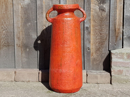 A large, red, textured vase with two handles stands against a wooden fence.