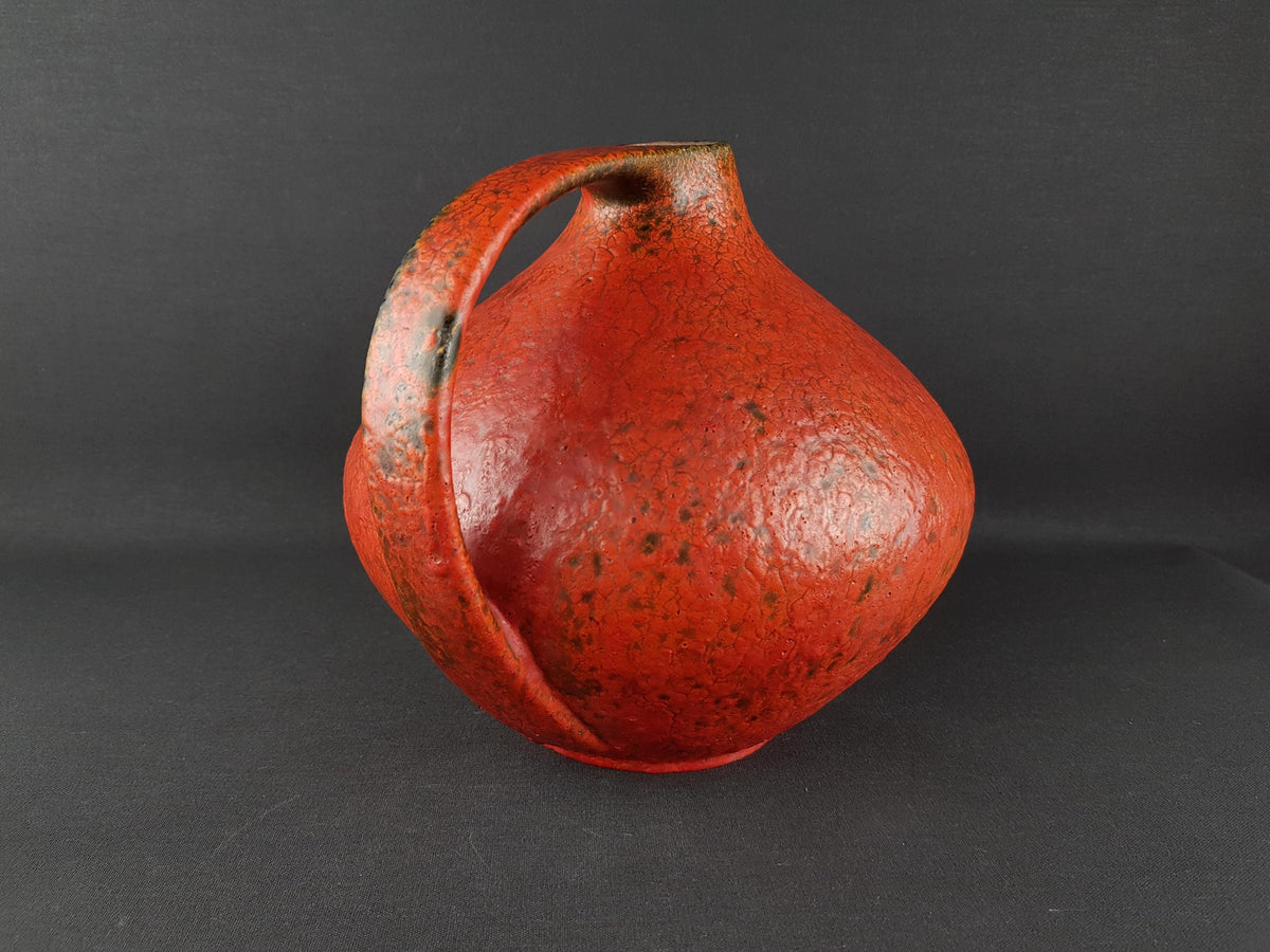 A red ceramic vase with a textured surface and a handle on one side, sitting on a dark surface.