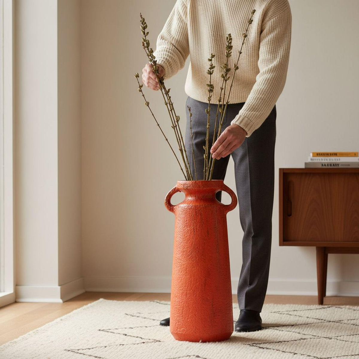 A person is arranging a bouquet of small branches in a tall, orange vase with handles on a white rug in a room.