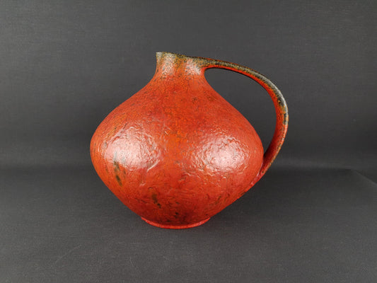 A red ceramic pitcher with a curved handle, sitting on a dark surface.