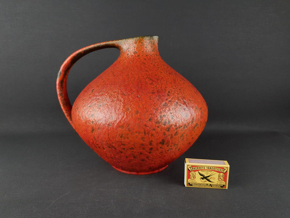 A large red ceramic pitcher with a curved handle, placed next to a matchbox on a dark background.