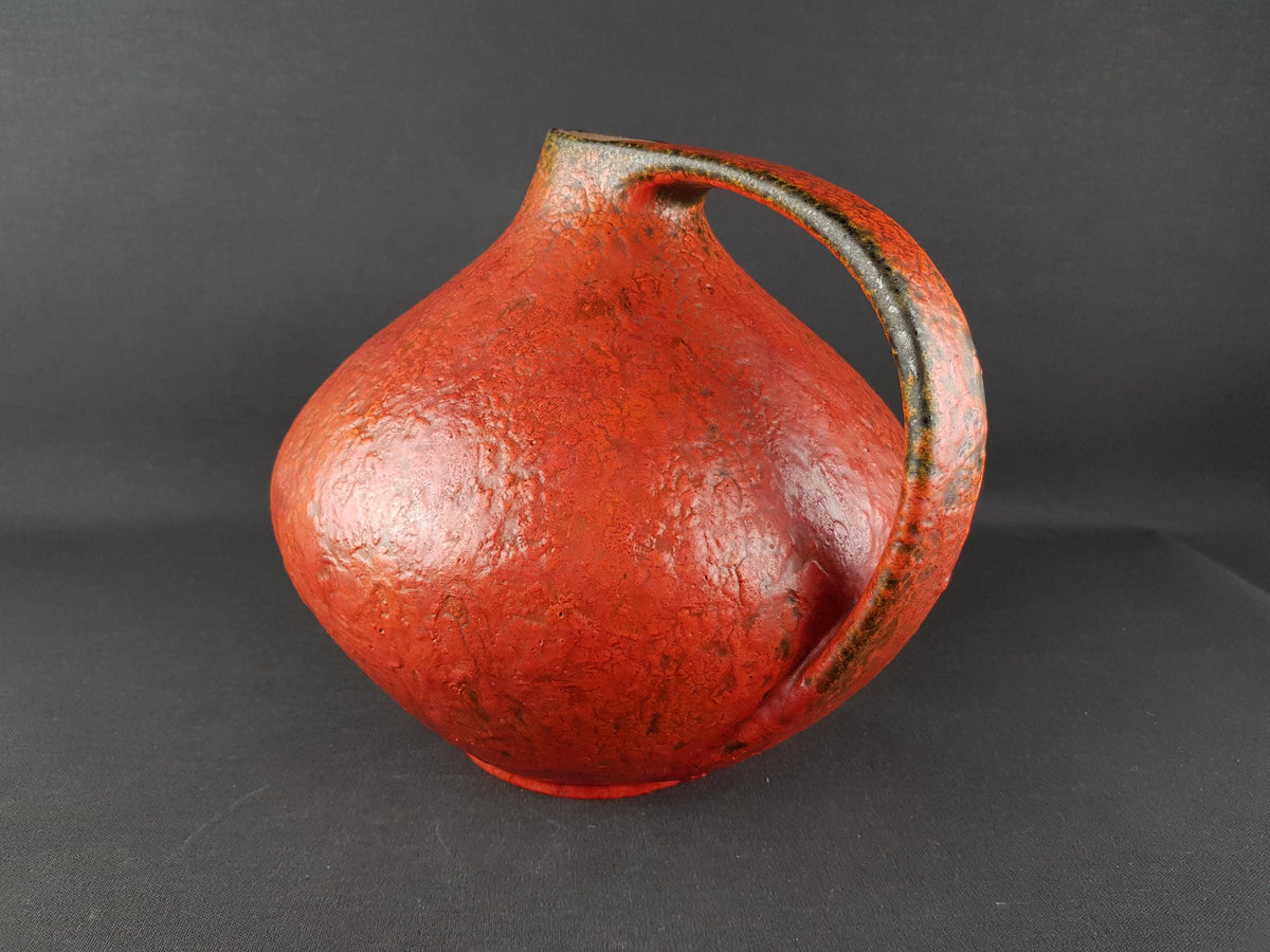 A red ceramic pitcher with a curved handle, sitting on a dark surface.