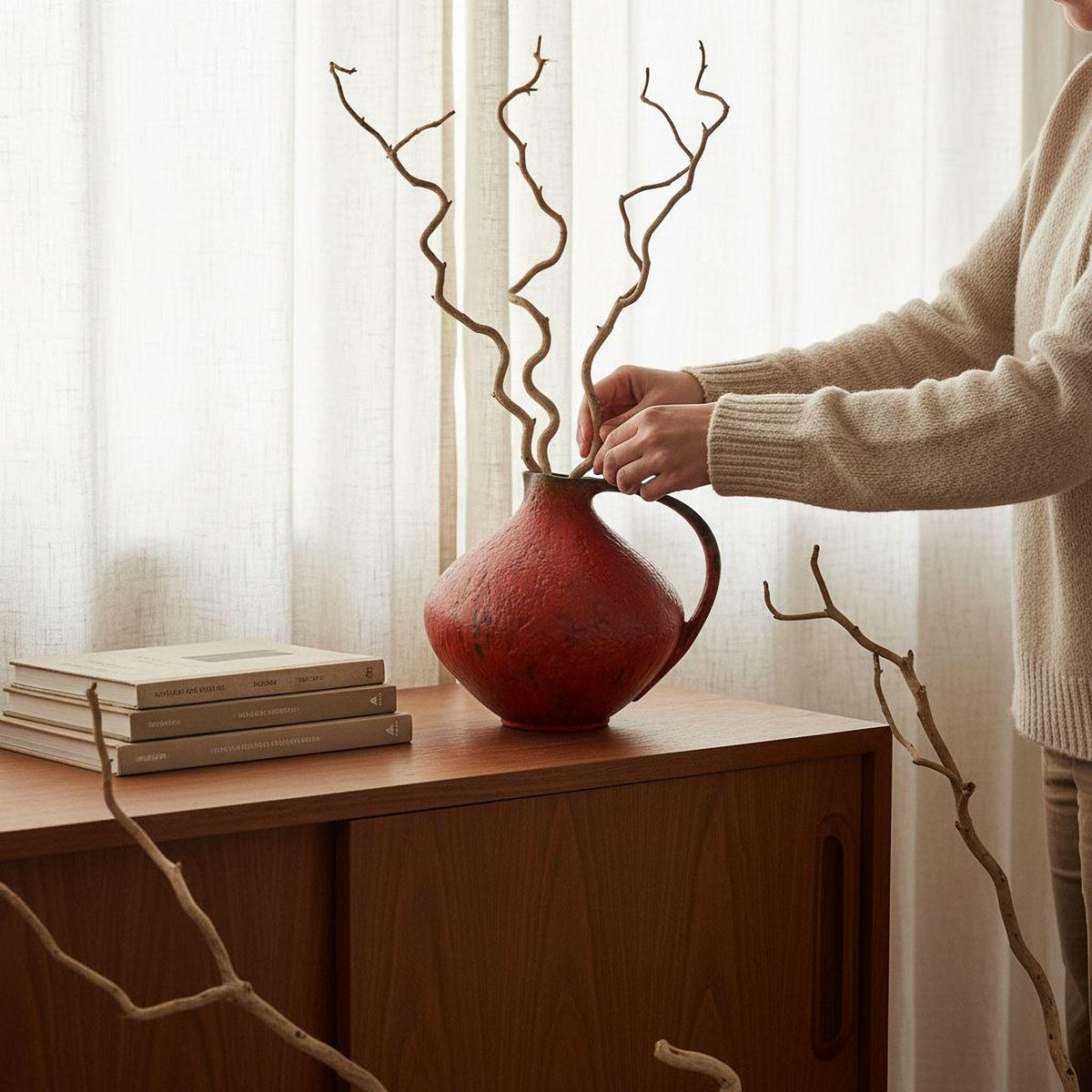 A person is arranging a red vase with twisted branches on a wooden table, surrounded by books and a stack of books.